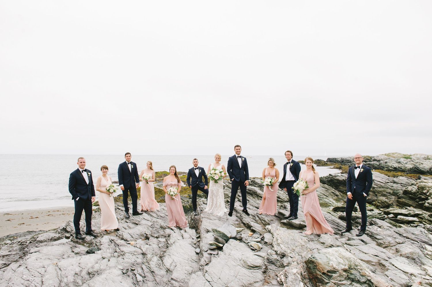Wedding party poses on rocky beach in blush pink bridesmaid gowns and navy tuxedos.