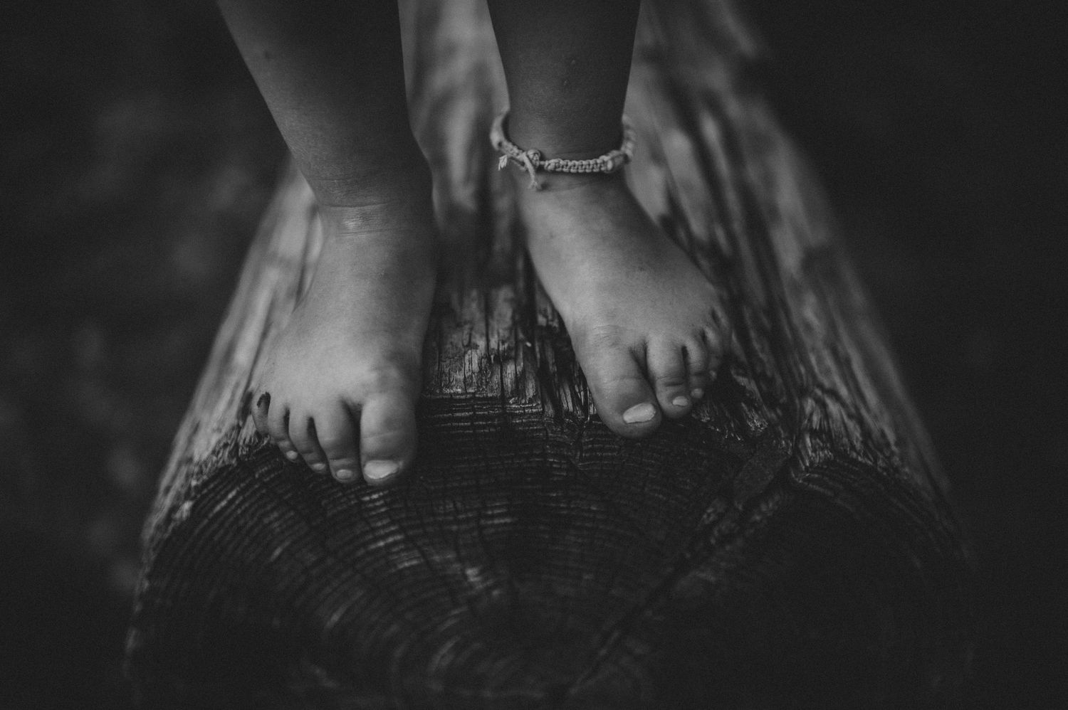 Black and white photo of a child's bare feet standing on a wooden log, wearing a delicate anklet bracelet.