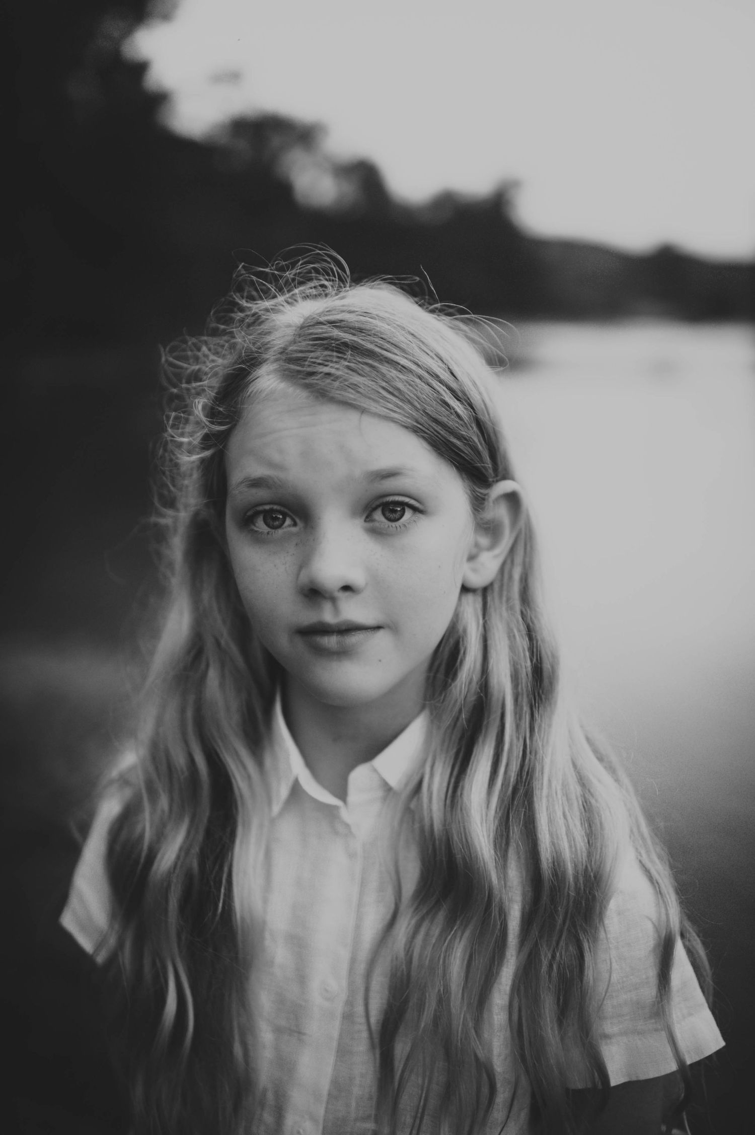 Black and white portrait of a young girl with long wavy hair outdoors near water.