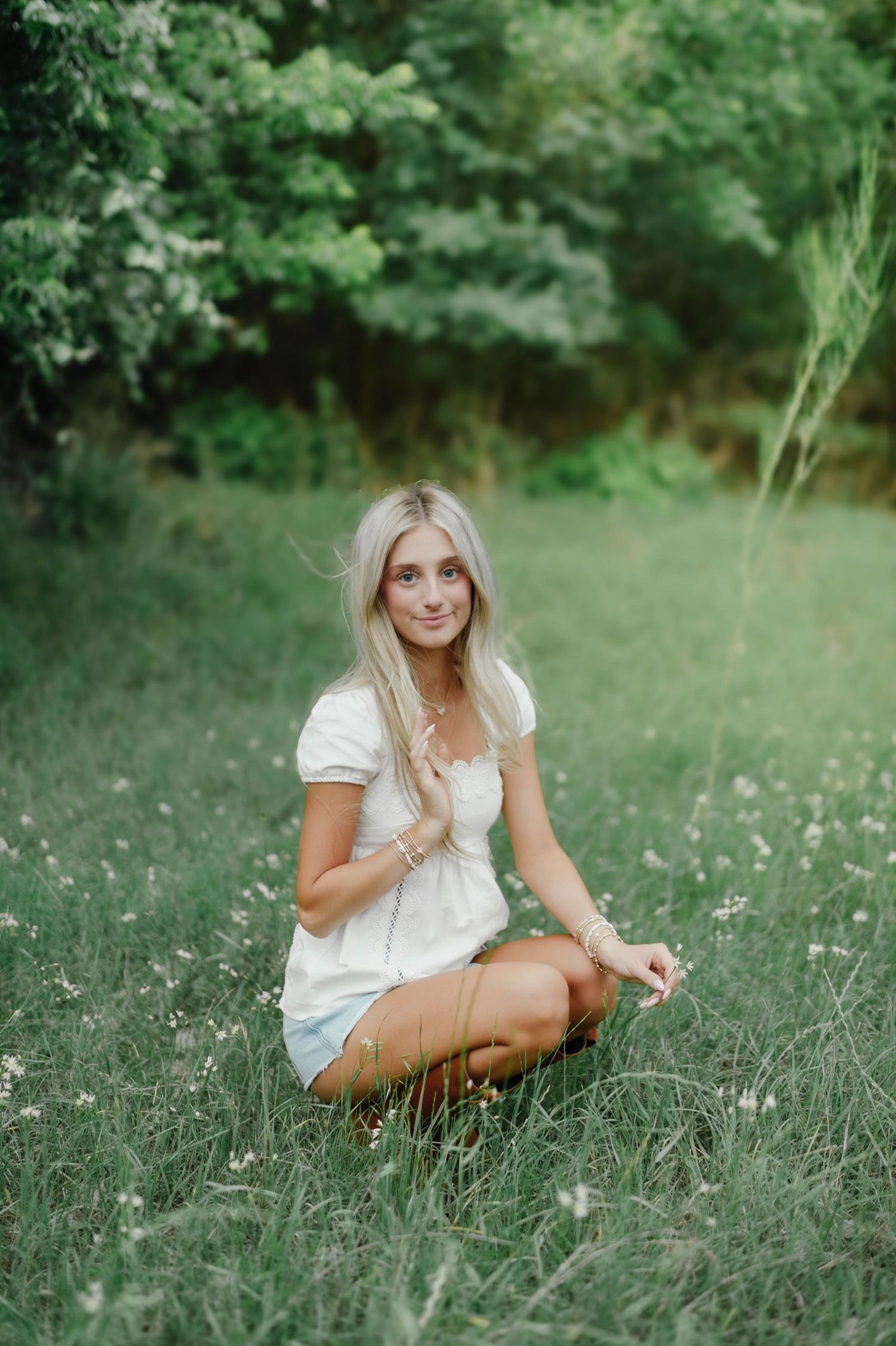 Young blonde woman in white top and denim shorts sitting cross-legged in a lush green meadow with wildflowers.