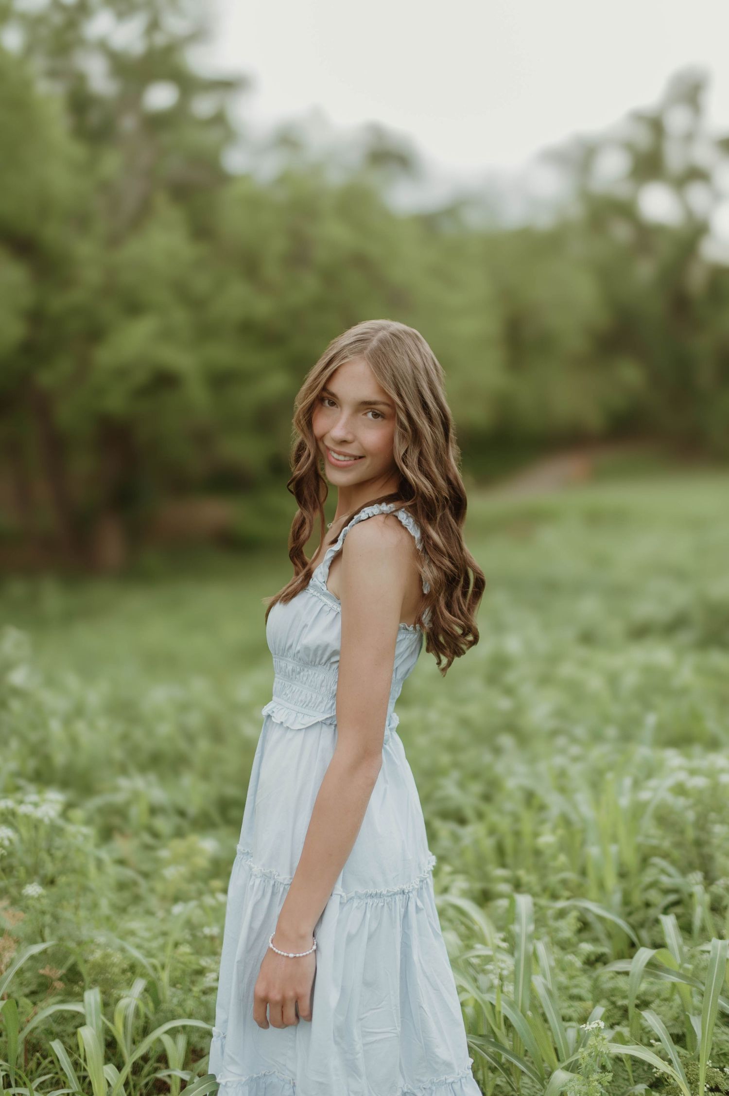 Young girl in light blue tiered maxi dress smiling in a lush green meadow during an outdoor portrait session.