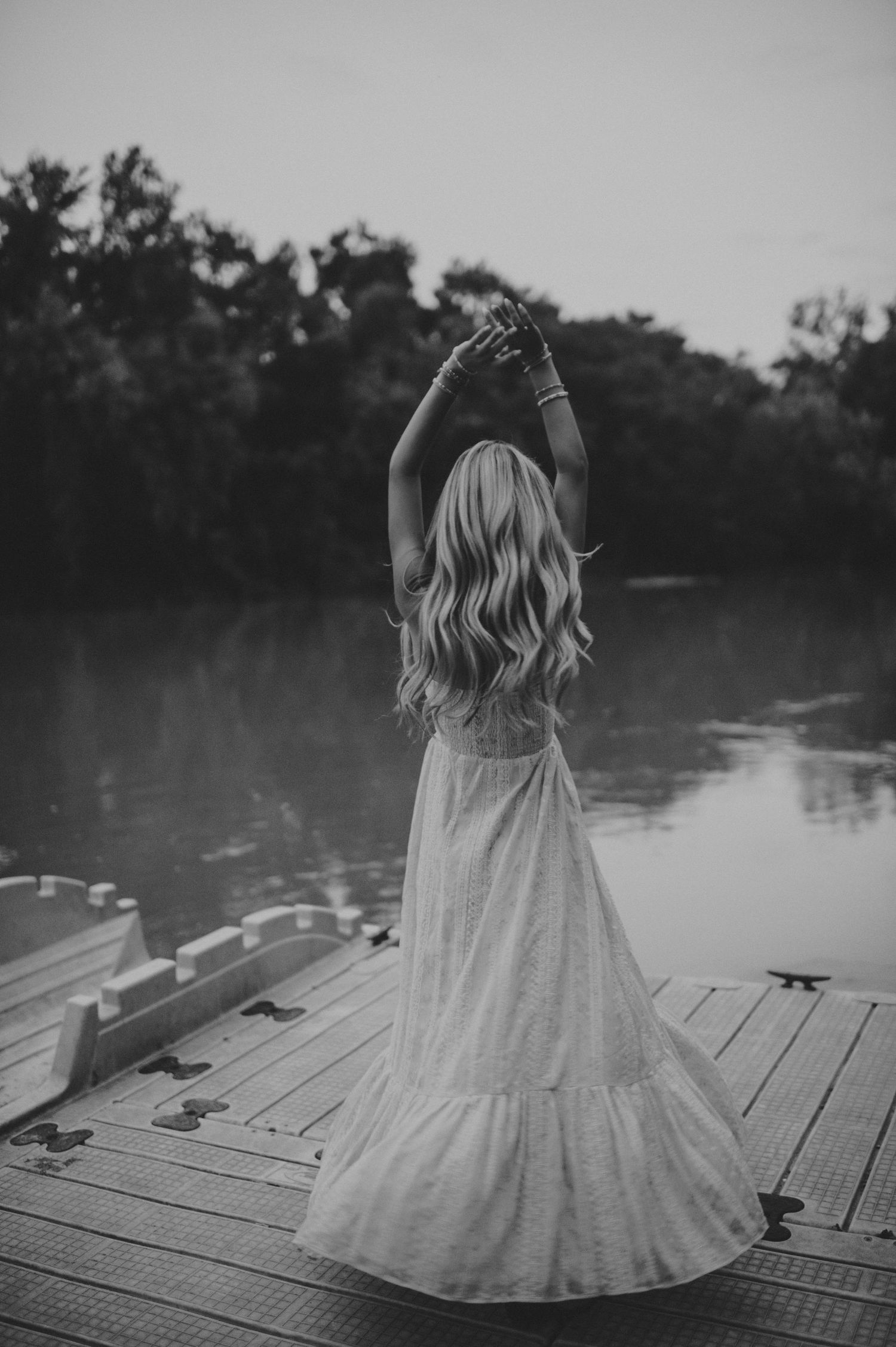 A blonde woman in a white maxi dress raises her arms on a wooden dock by a serene lake.