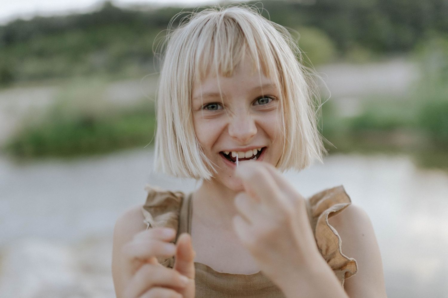 Laughing blonde girl with bangs eating a snack outdoors near a river.