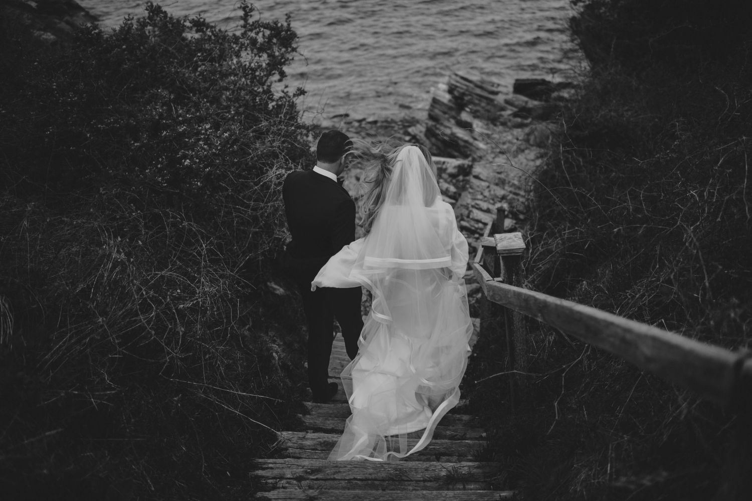 Bride and groom walk down wooden steps toward the ocean in a romantic black and white wedding photo.