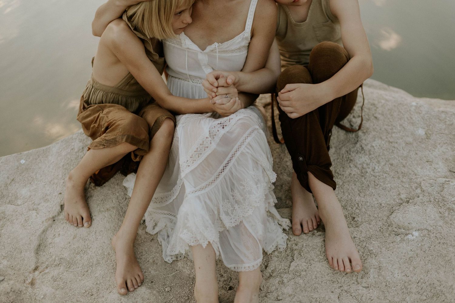 A woman in a white dress holds hands with two children while sitting together on a rock.