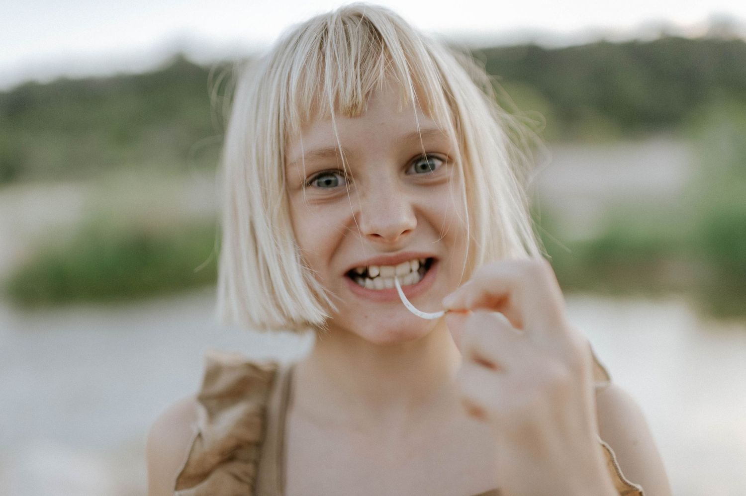 Cheerful blonde girl with bangs enjoying a treat near a blurred outdoor background.