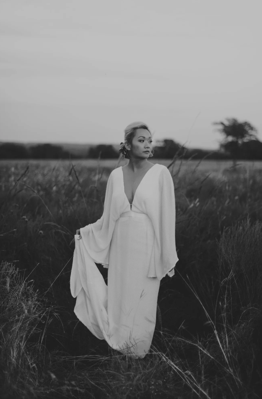 Black and white photo of bride in flowing white dress with wide sleeves standing in a field at dusk.