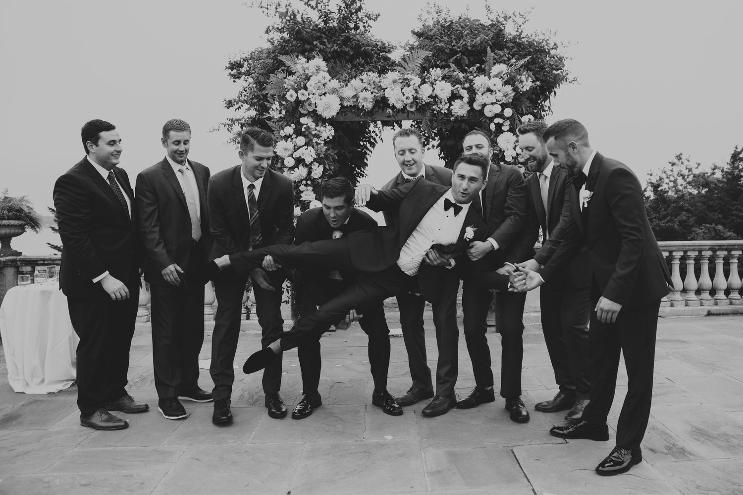 Black and white photo of groomsmen playfully lifting groom during fun outdoor wedding party celebration.