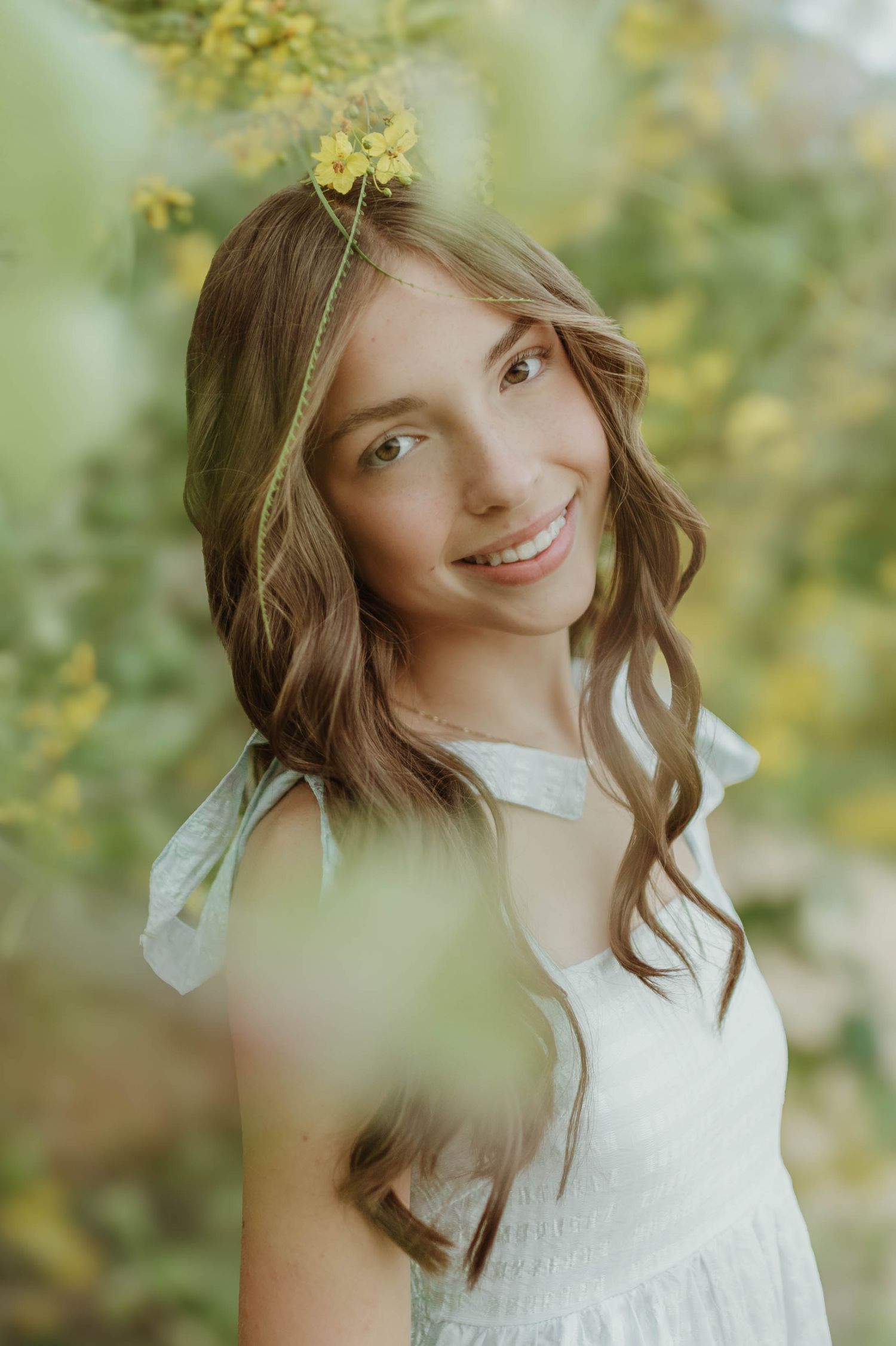 Smiling teenage girl in white dress surrounded by yellow blooms in a soft-focus outdoor portrait.
