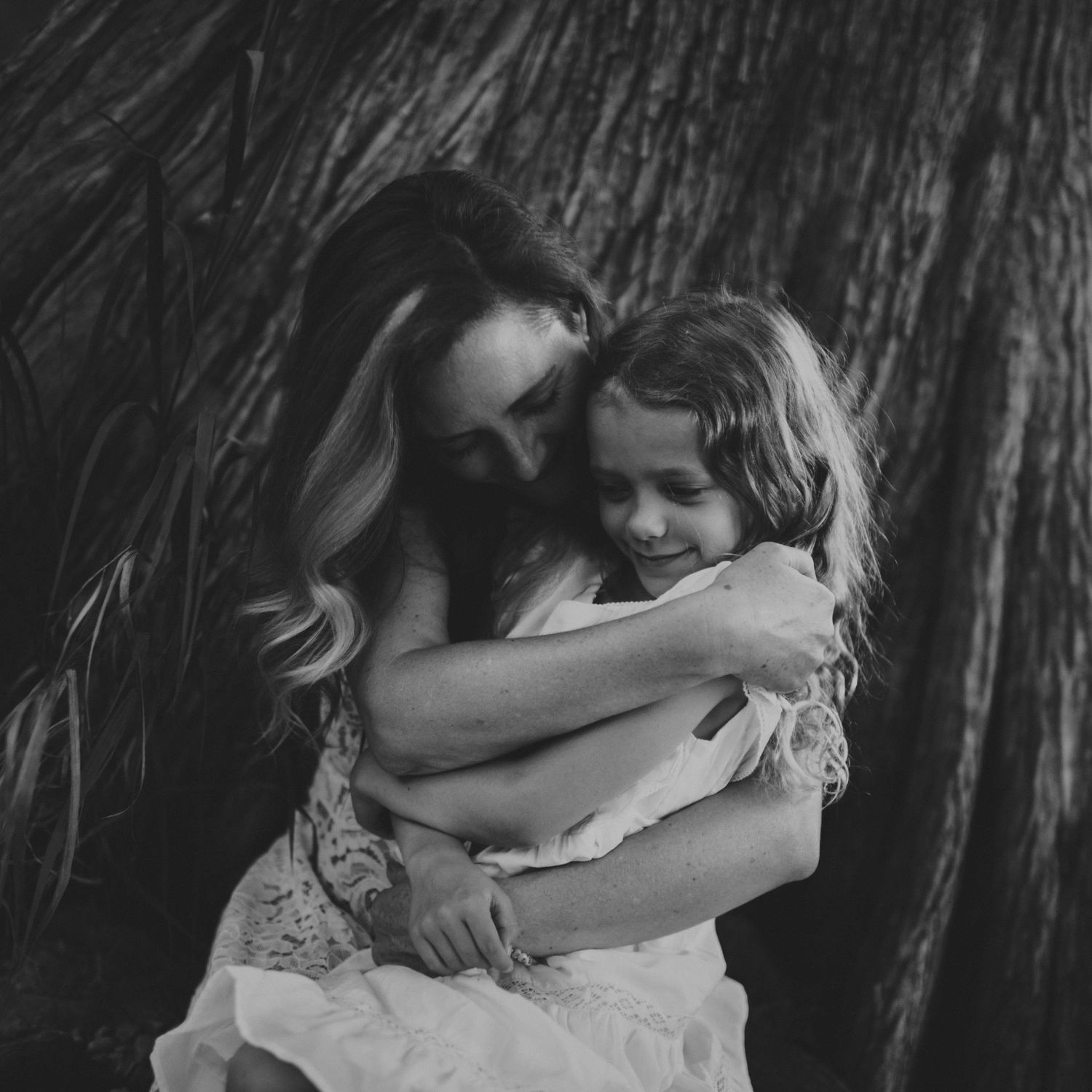 A mother tenderly embracing her young daughter against a rustic tree backdrop in black and white.