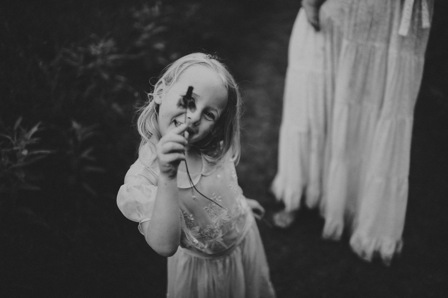 Black and white photo of a young blonde girl in a white dress holding a flower outdoors.