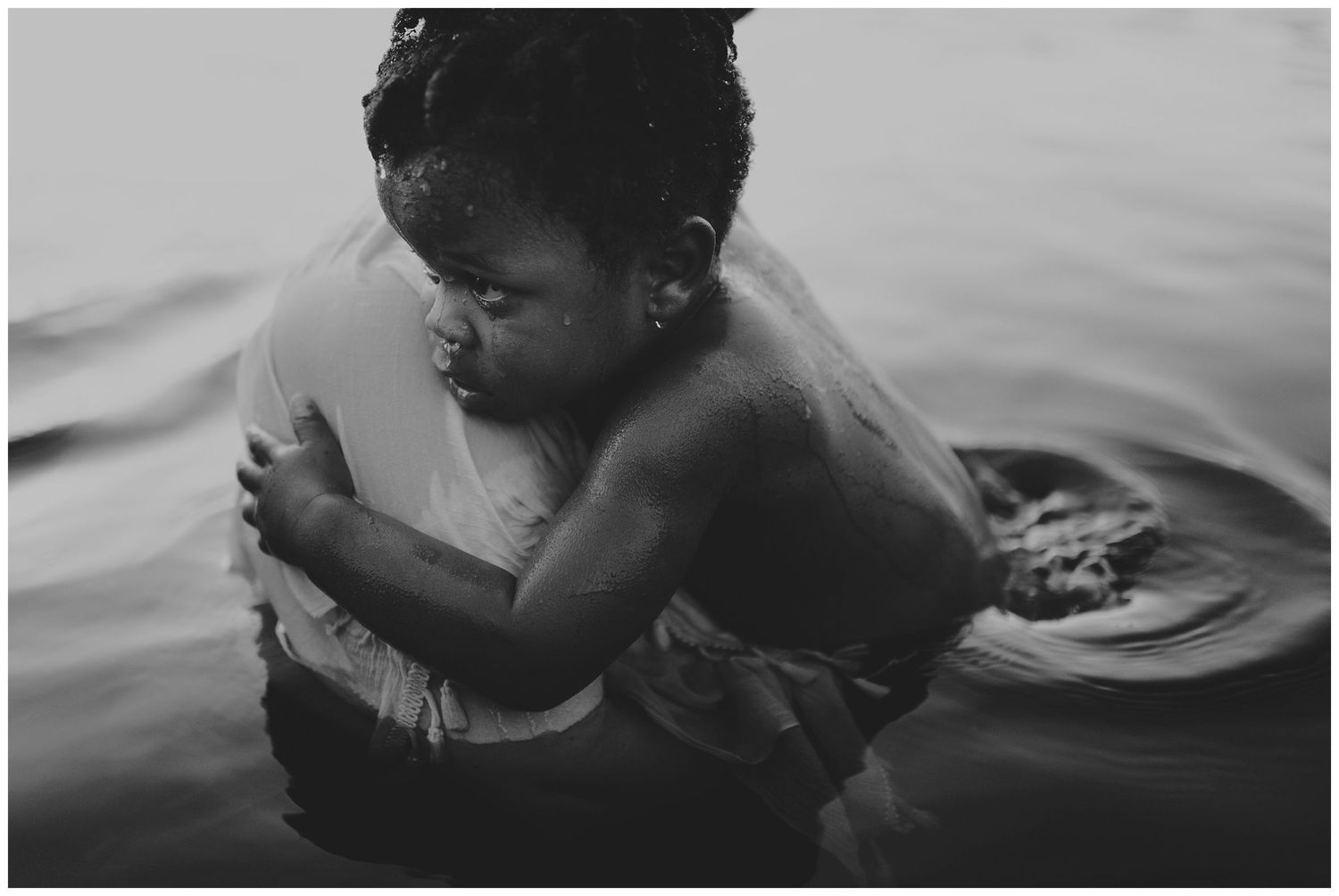 Black and white photo of a young child hugging an adult while standing in calm water.
