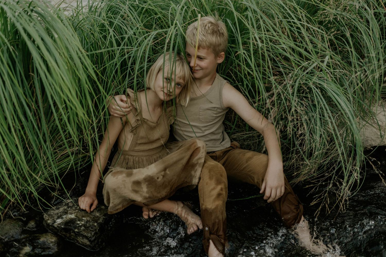 Two blonde children sit together on rocks beside a stream surrounded by tall green grass.