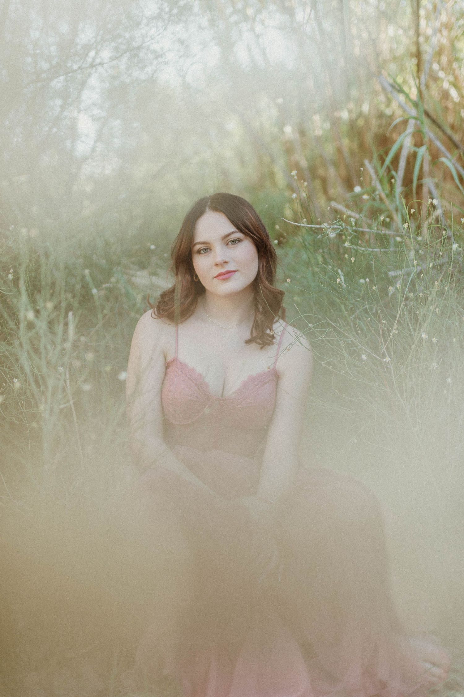Young woman in pink dress sitting among soft wildgrasses in dreamy outdoor portrait.