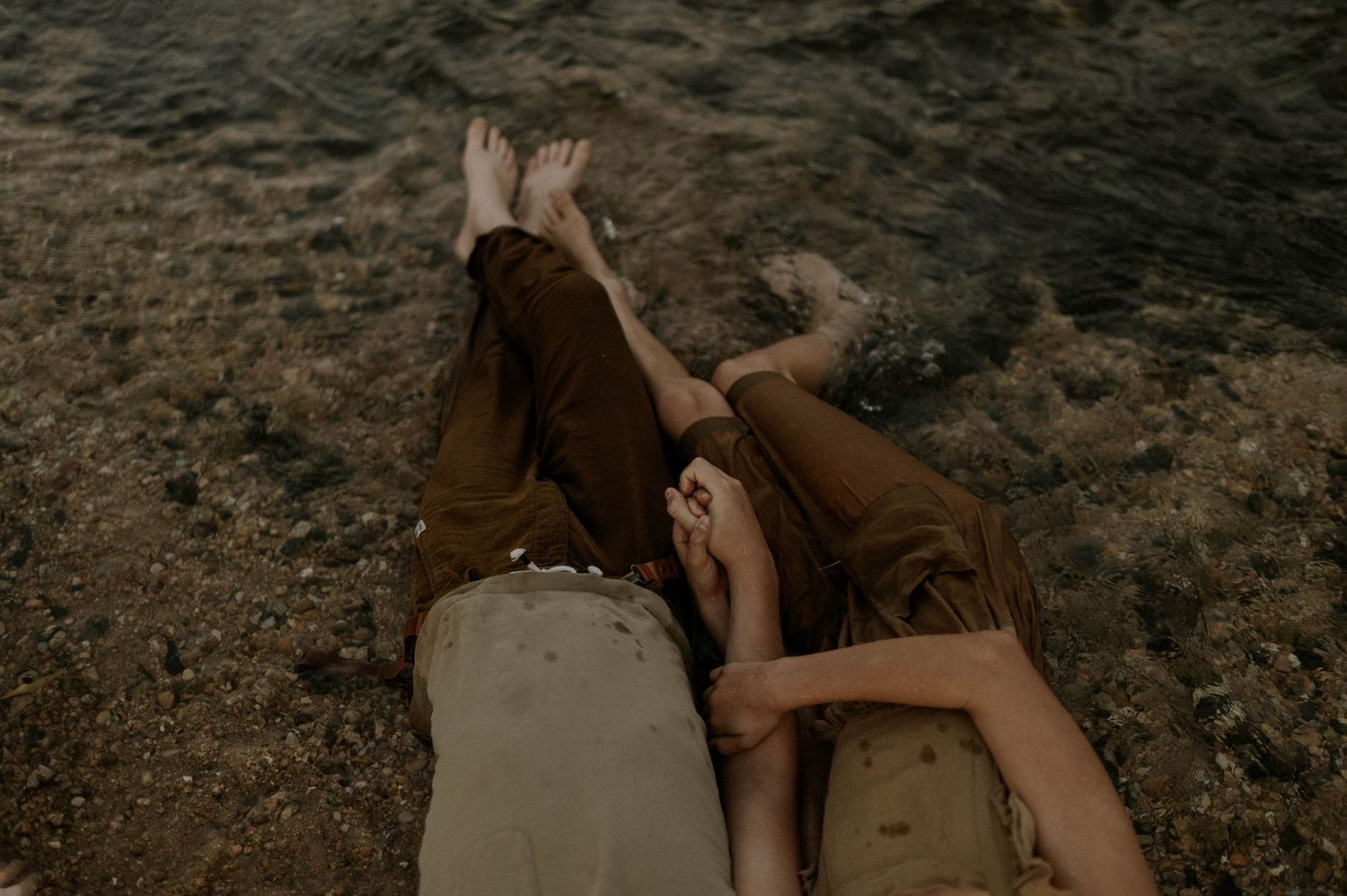 Two children lying on sandy shore holding hands with feet near water.