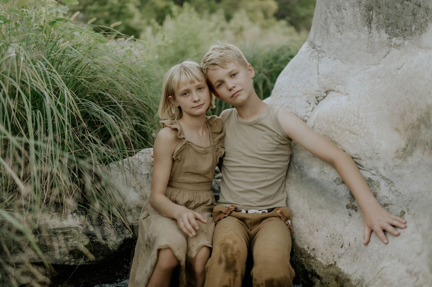 Two blonde children in earthy tones sit together smiling beside a large rock outdoors.