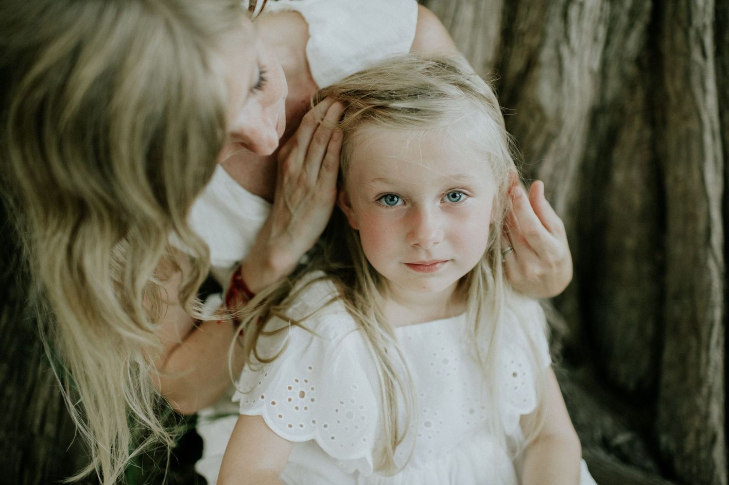 A woman gently fixes the hair of a young blonde girl in a white dress, both with soft natural lighting.
