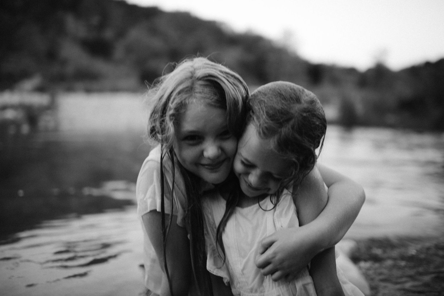 Two young girls hugging and smiling near a river in a black and white outdoor portrait photo.