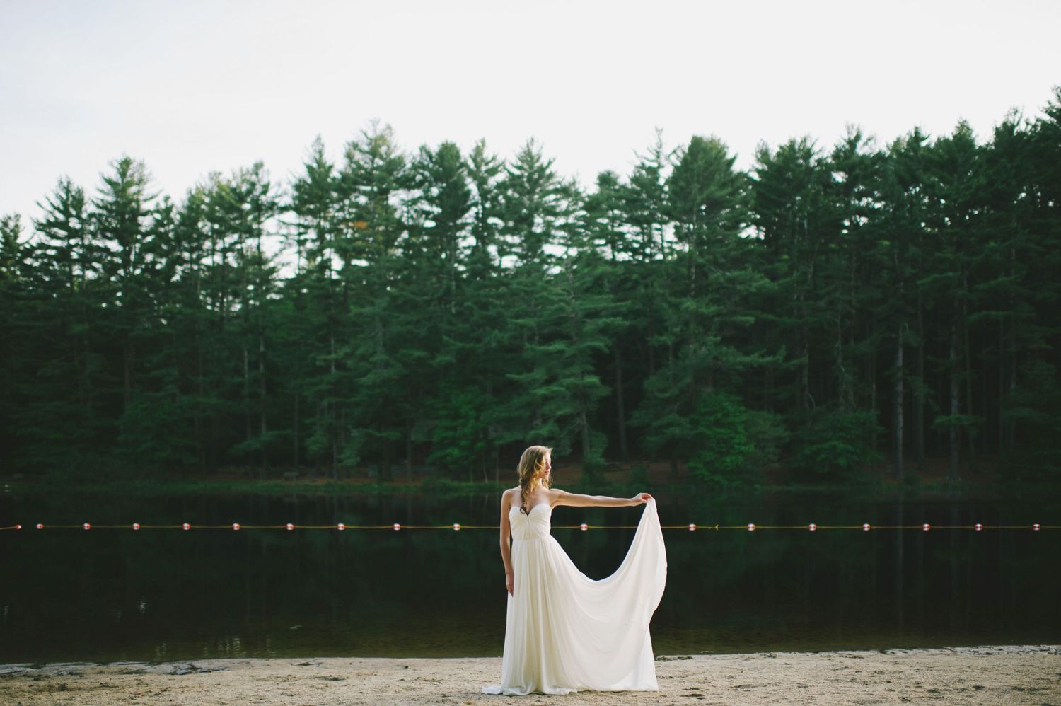 Bride in flowing white gown spreads cape wide while standing on sandy shore before pine forest lake.