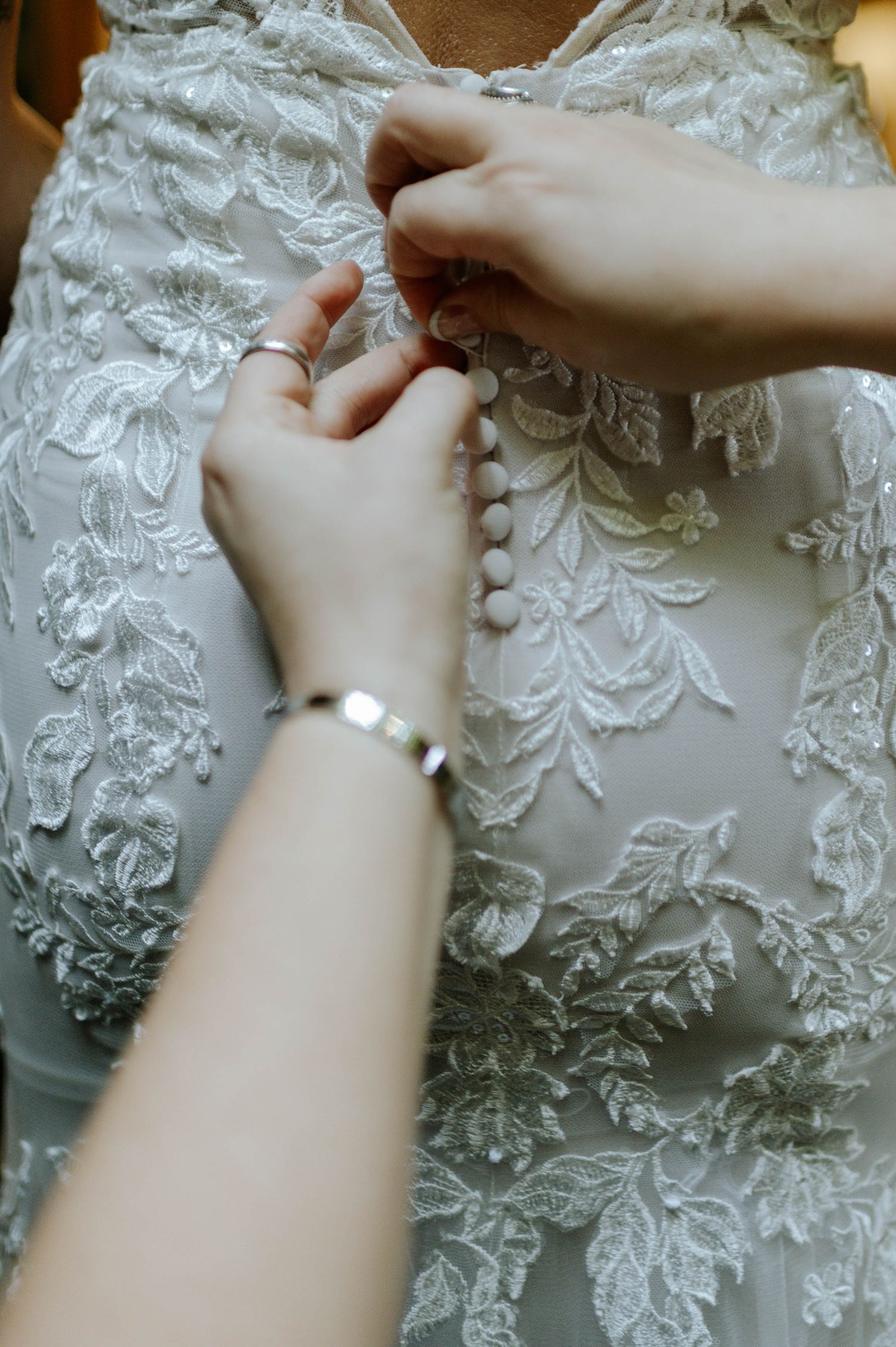 Close-up of hands buttoning pearl buttons on a lace wedding dress with a bracelet visible.