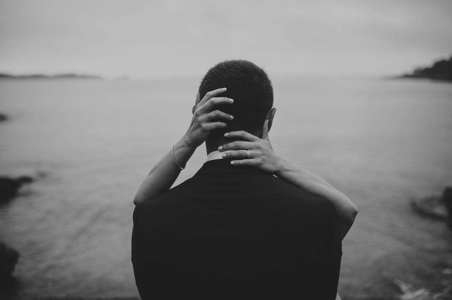 Black and white rear view of person clasping hands behind neck while overlooking misty ocean landscape.