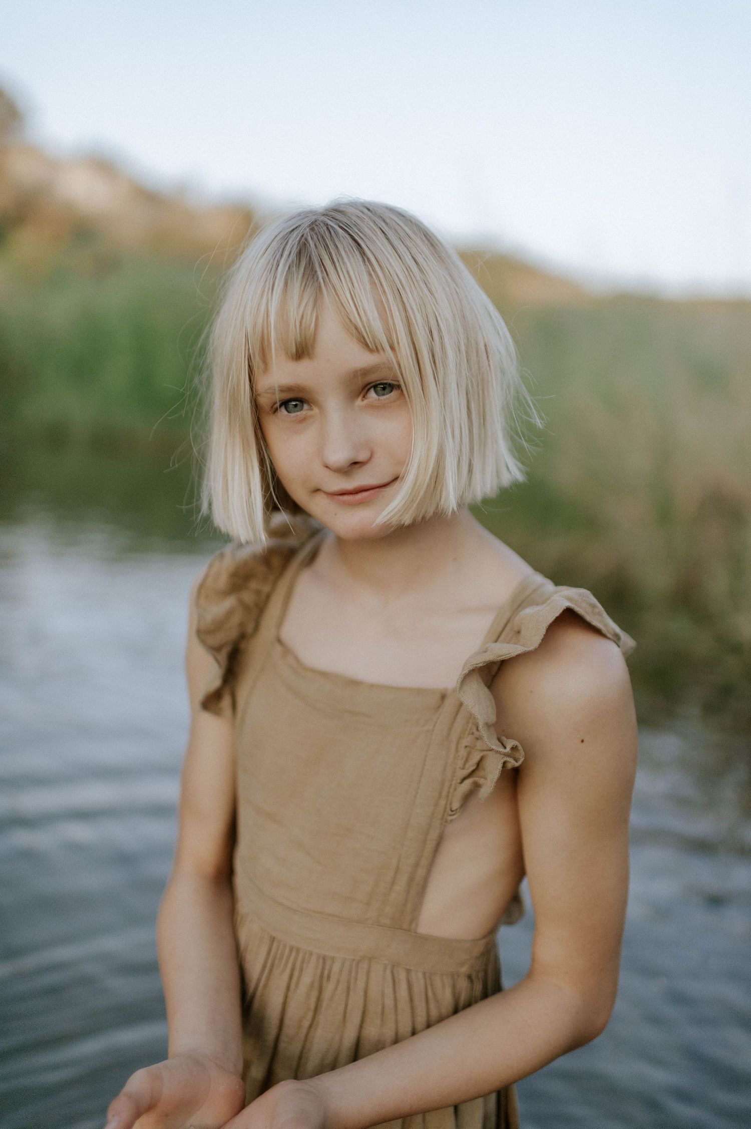 Portrait of a blonde girl in a golden sundress standing near a peaceful river.