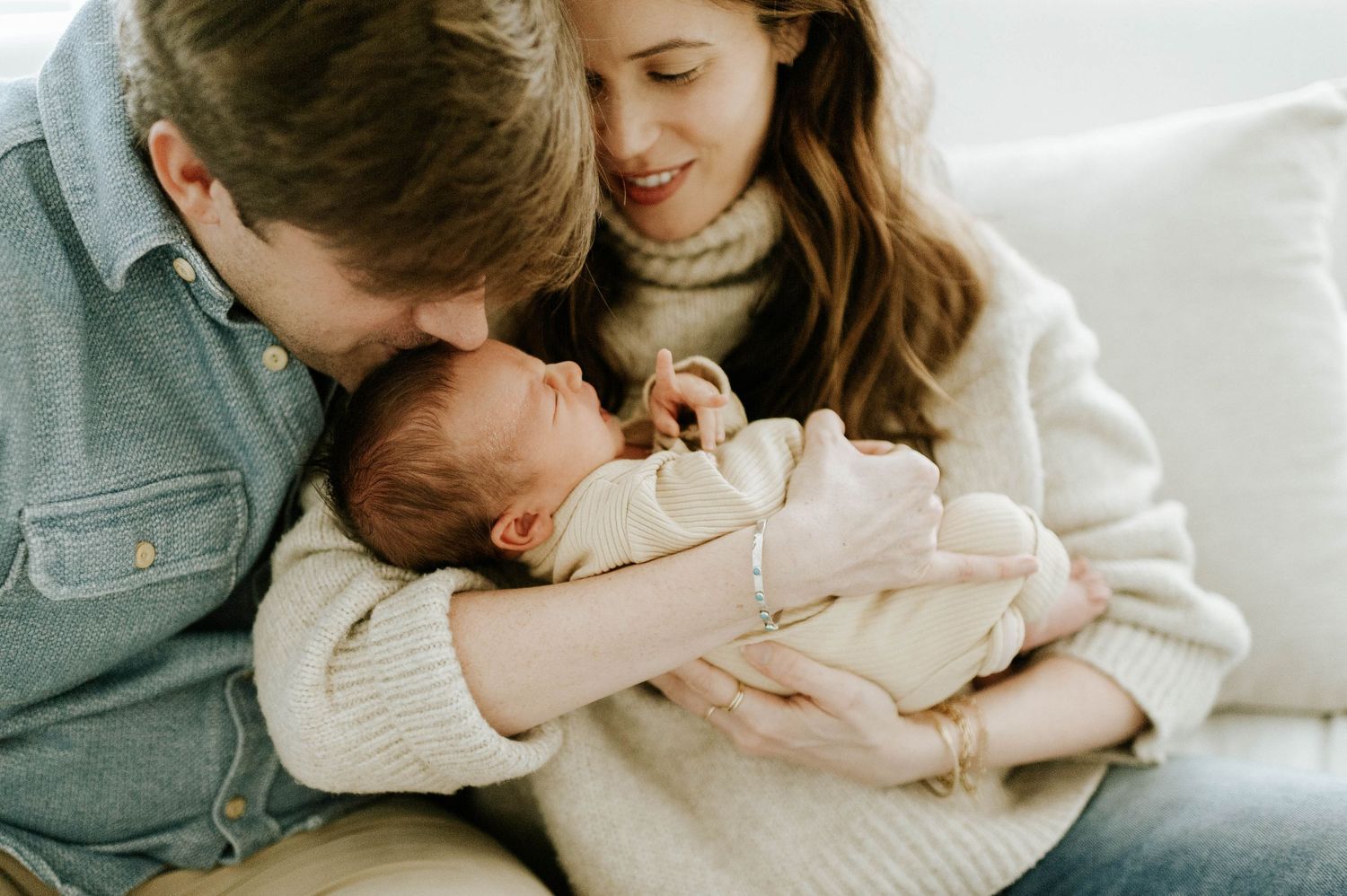Young parents tenderly kissing their newborn baby while sitting together on a white couch.