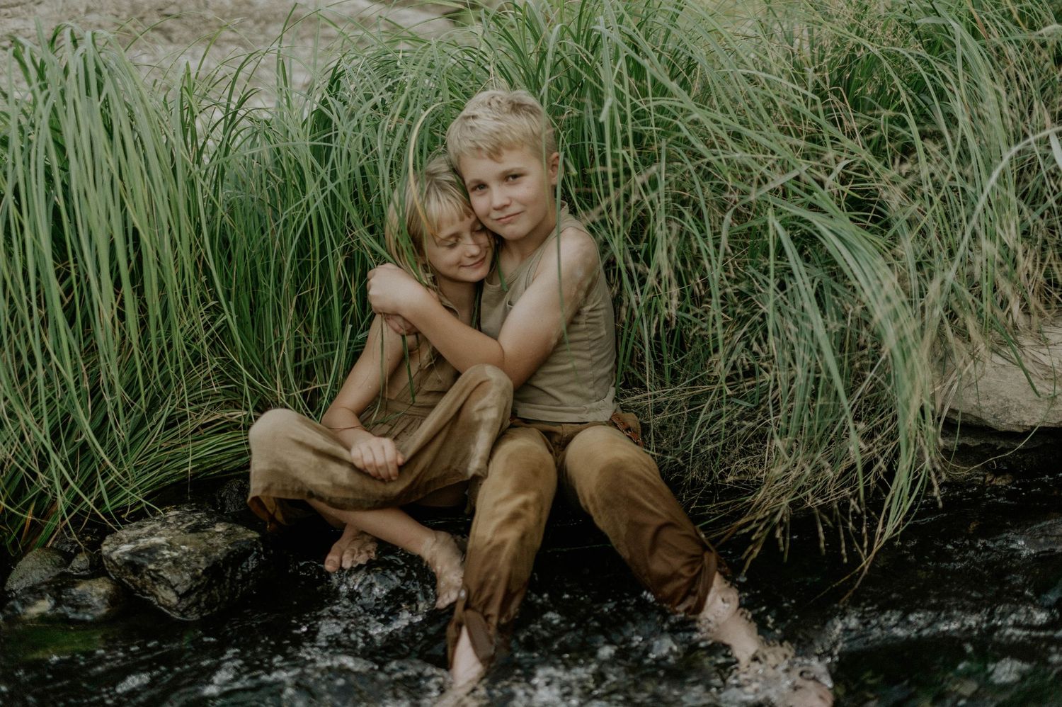 Two children sit together hugging by a flowing stream surrounded by tall green grass outdoors.