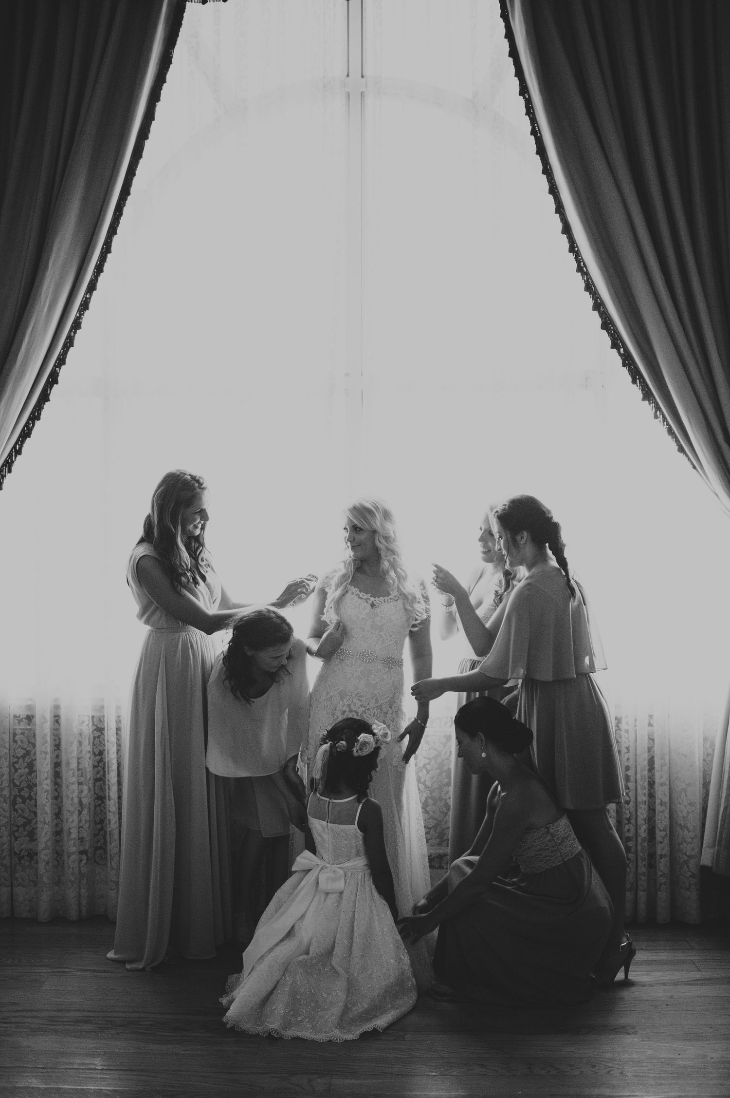 Bride surrounded by bridesmaids and flower girl in front of a bright window.