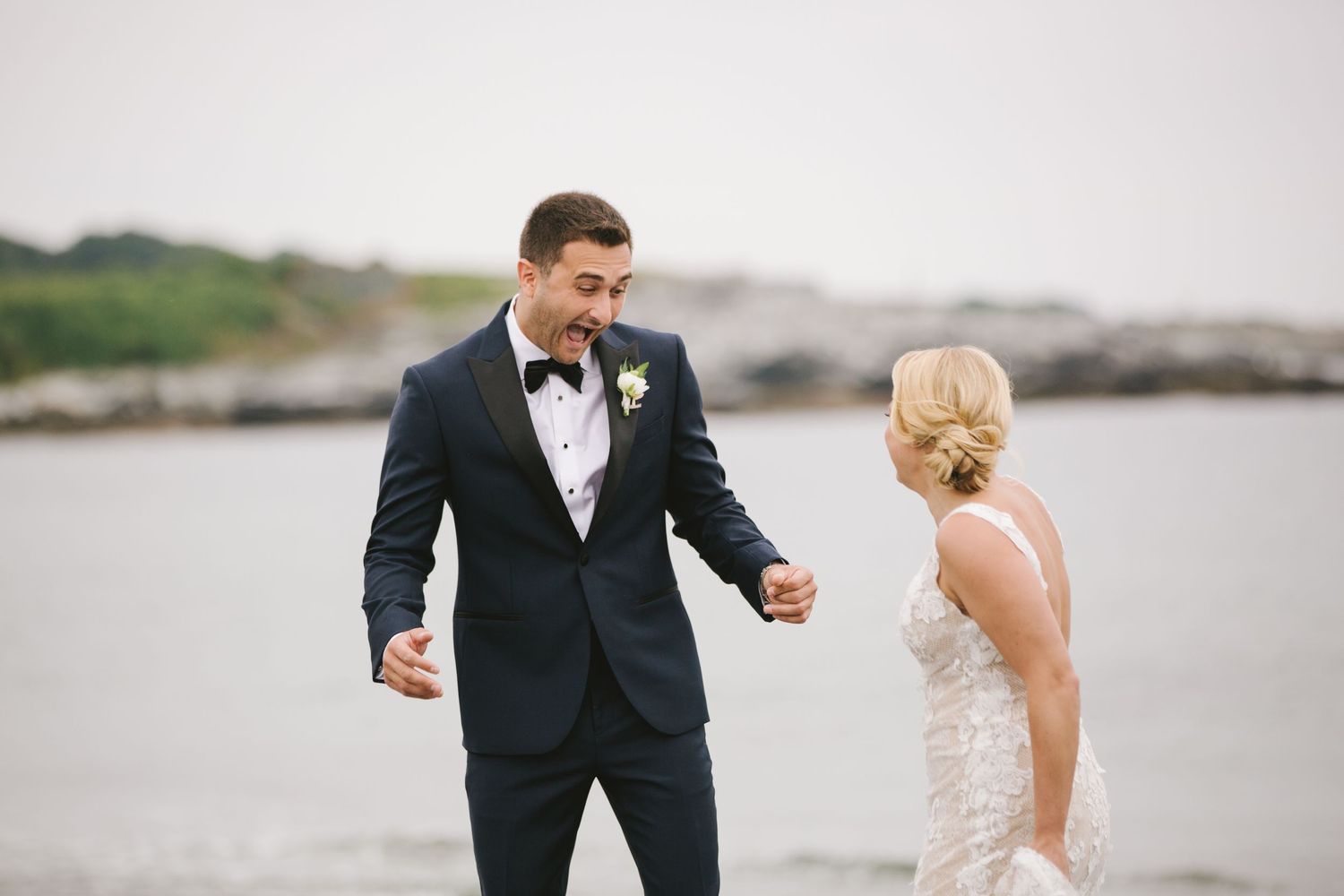 Groom in navy tuxedo reacts joyfully seeing bride during first look by the ocean.