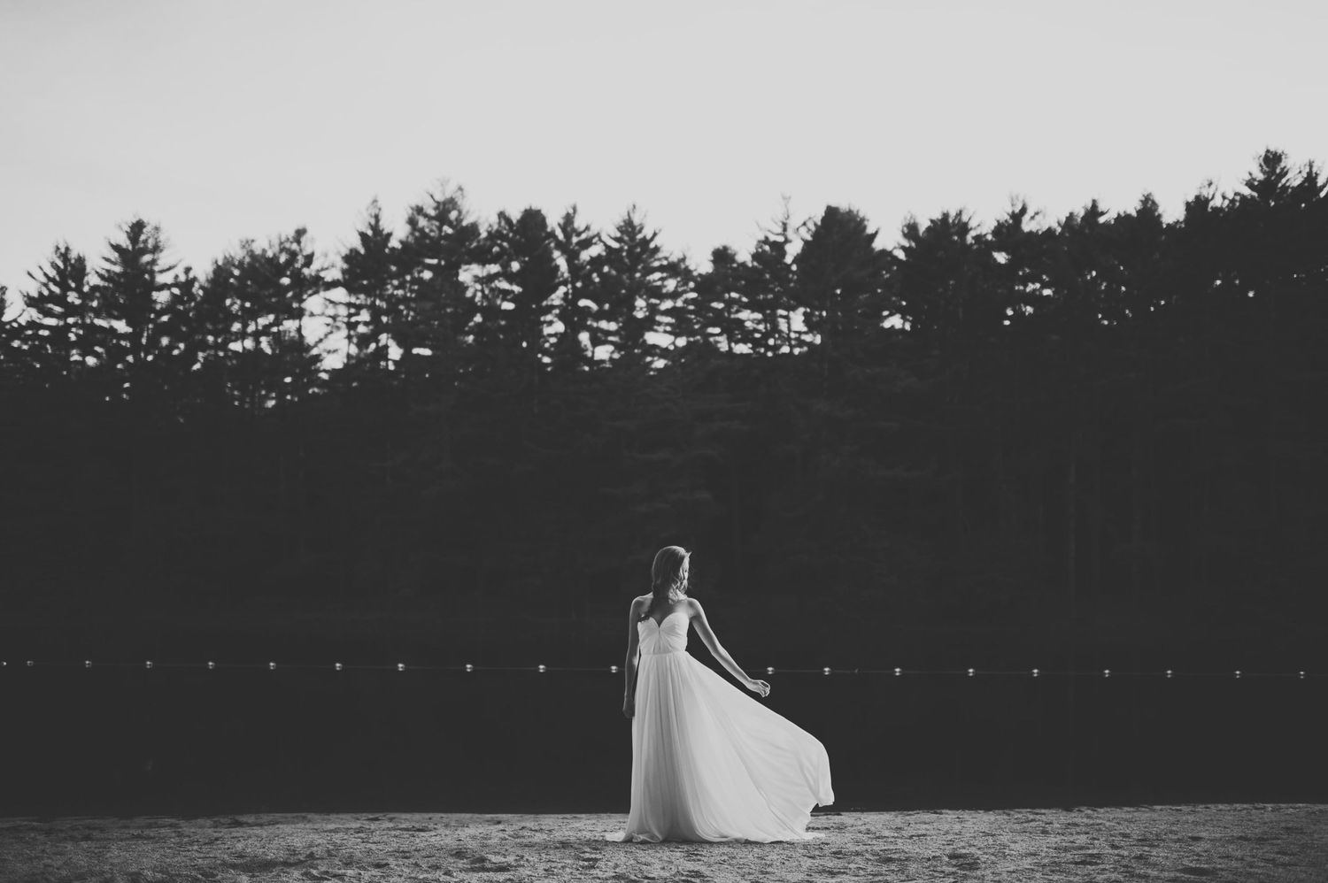 Black and white photo of bride in flowing white dress standing on beach near forest at dusk.