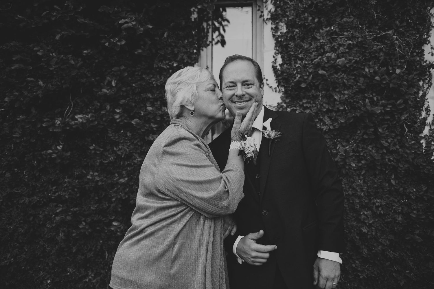 Elderly woman kisses groom on the cheek outdoors near ivy-covered wall.
