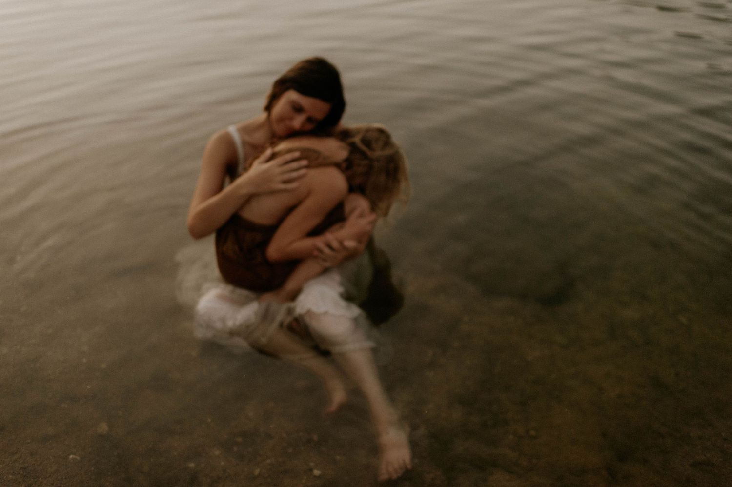 Two women hug closely while sitting in serene water, one in white dress and one in brown.