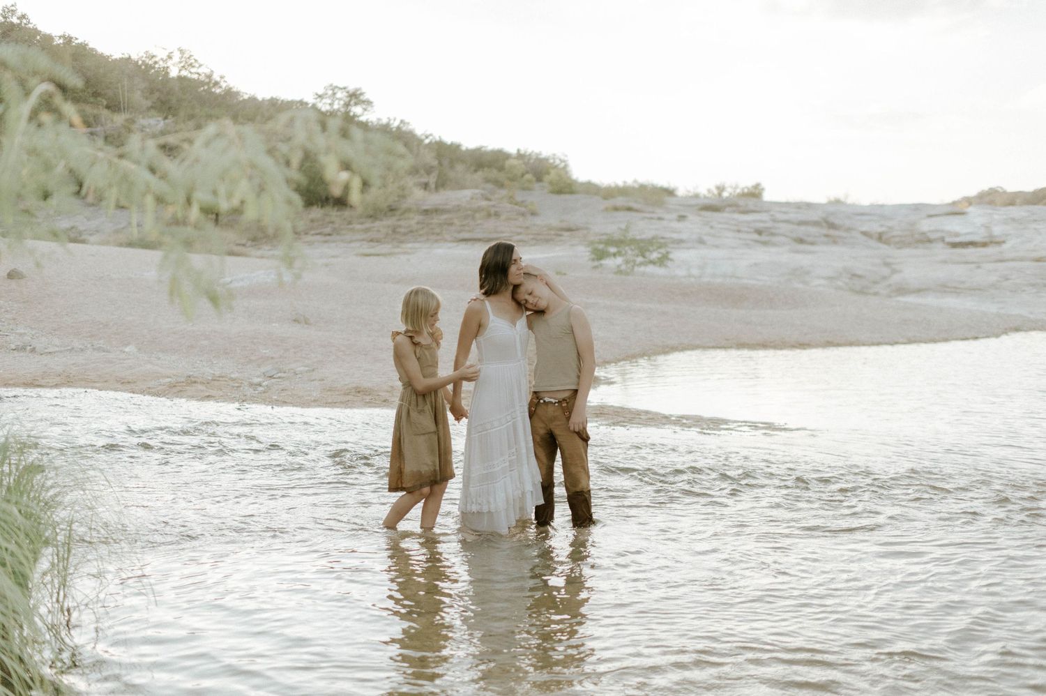 A family of three stands together in shallow water near a sandy riverbank at dusk.