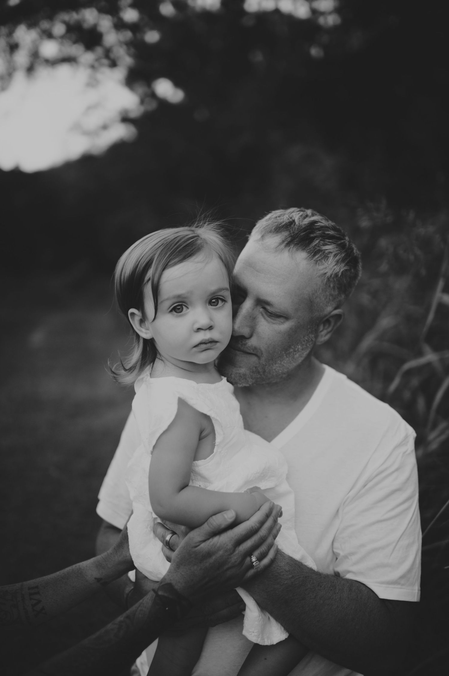 Black and white photo of a bearded man kissing a toddler girl in a white dress outdoors.