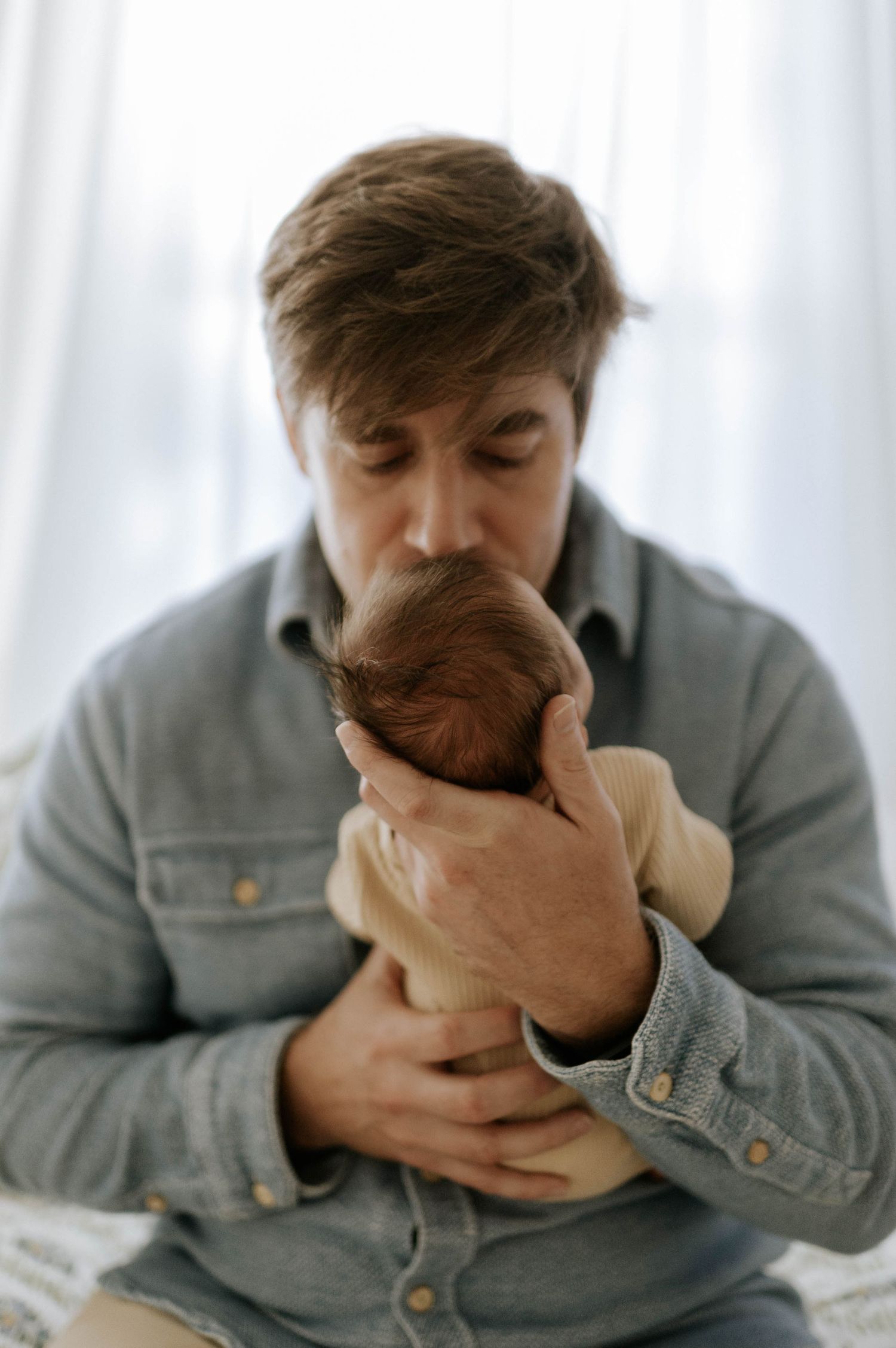 A father tenderly kisses his sleeping newborn baby, cradling the infant gently in both hands.