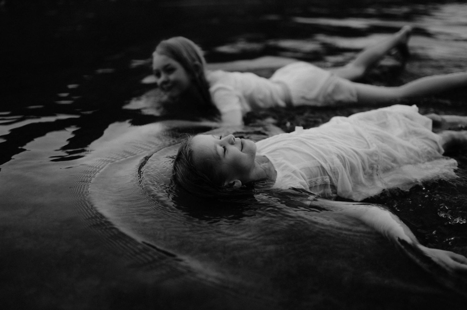 Two women in white dresses floating peacefully in dark water, captured in moody black and white photography.