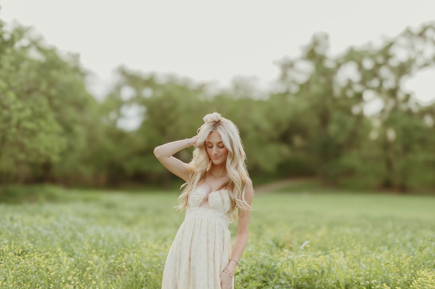 Blonde woman in cream dress standing in green meadow with hand in hair in soft natural light.