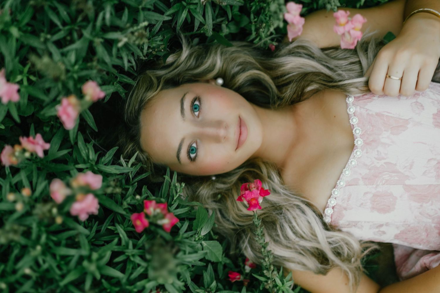 Young woman with blue eyes and wavy blonde hair lying among green foliage and pink flowers in a serene outdoor setting.