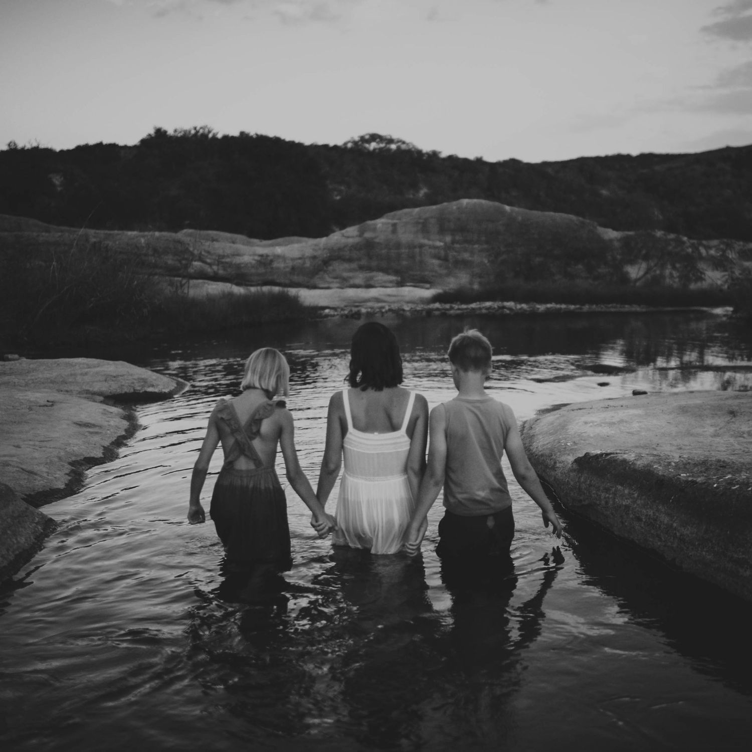 Three people holding hands walking through a shallow river toward scenic hills, black and white.
