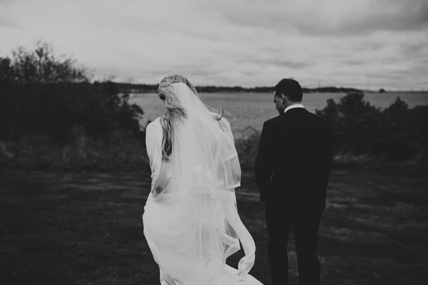 Black and white photo of a bride and groom walking through an open field with her veil flowing.