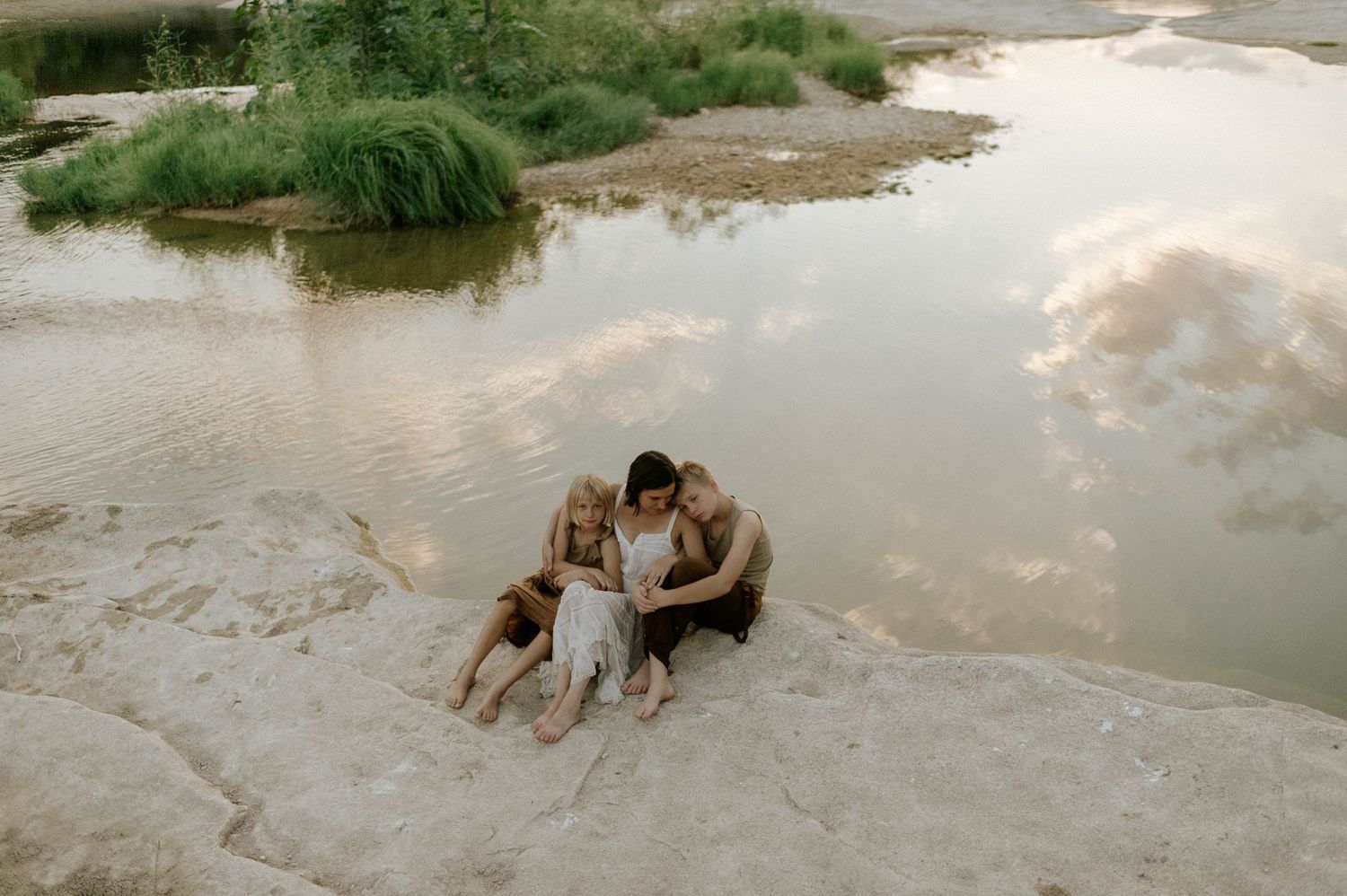 Family of three sitting together on rocky riverbank surrounded by lush greenery and calm water.
