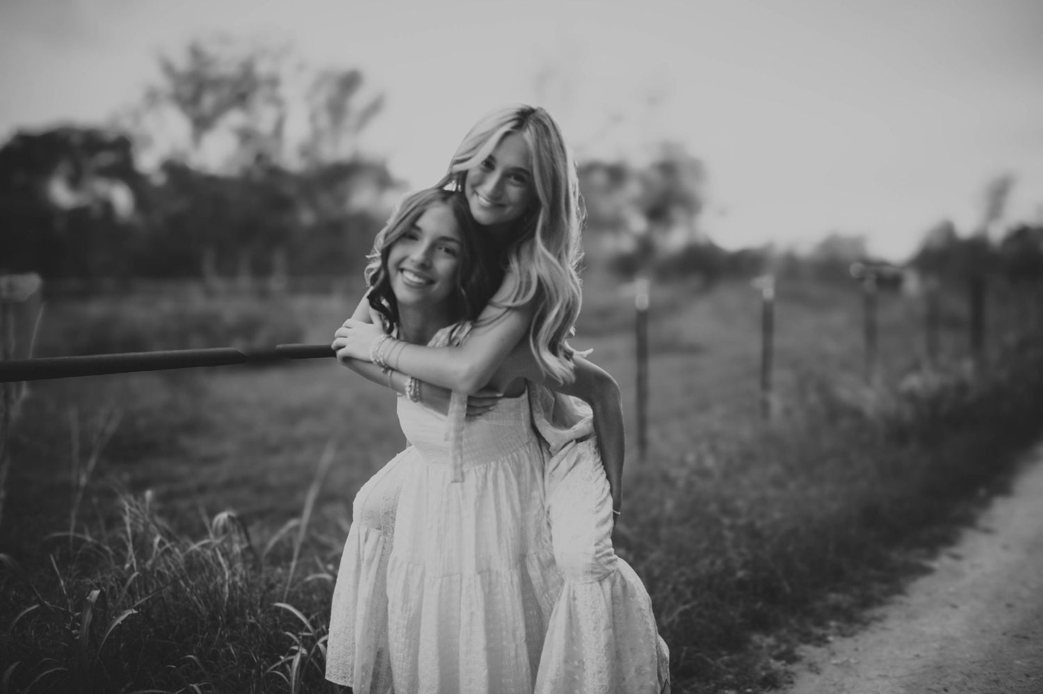 Two girls in white dresses laughing and hugging by a wooden fence in a charming black and white photo.