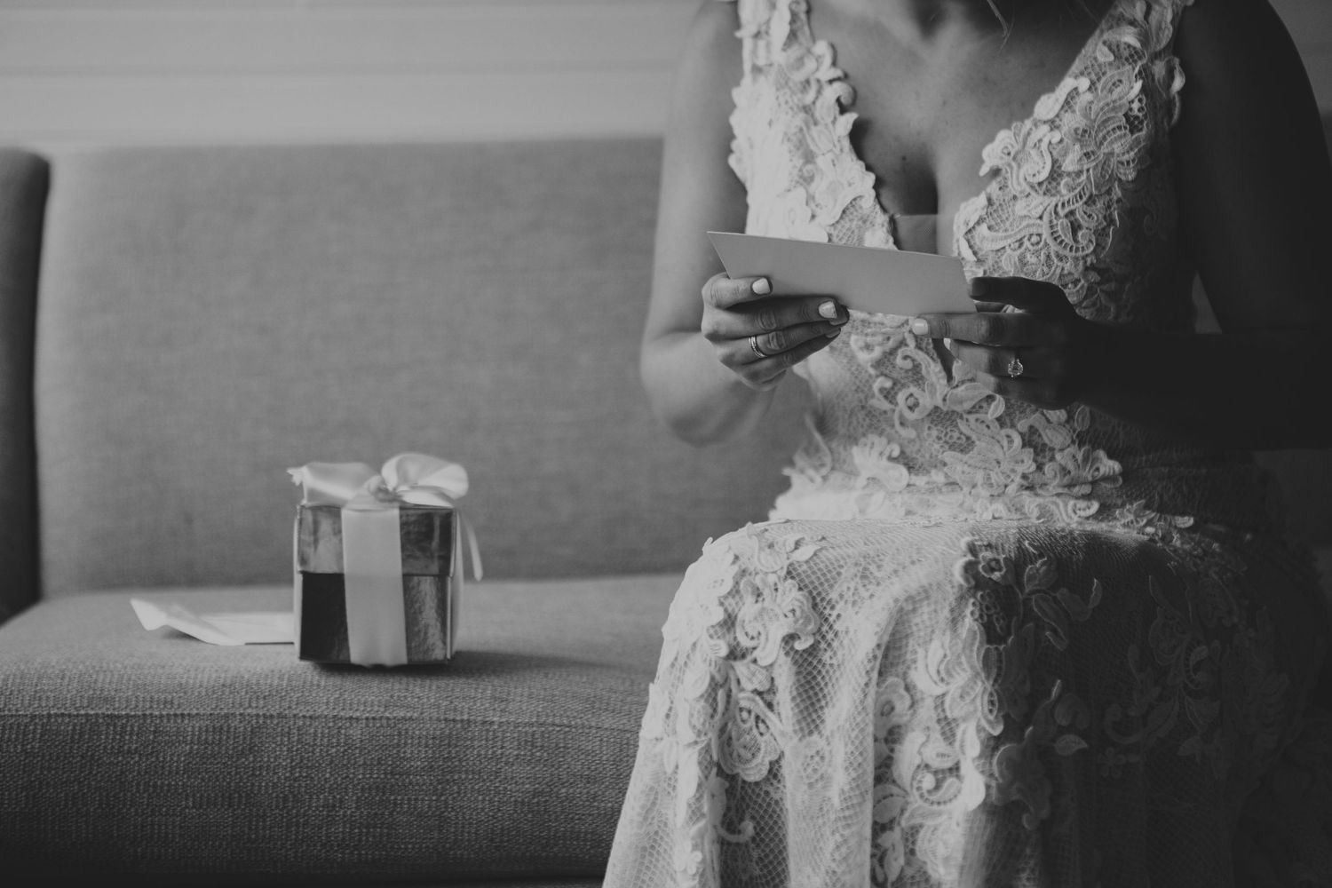Bride in lace dress reads a heartfelt note while sitting on a sofa.