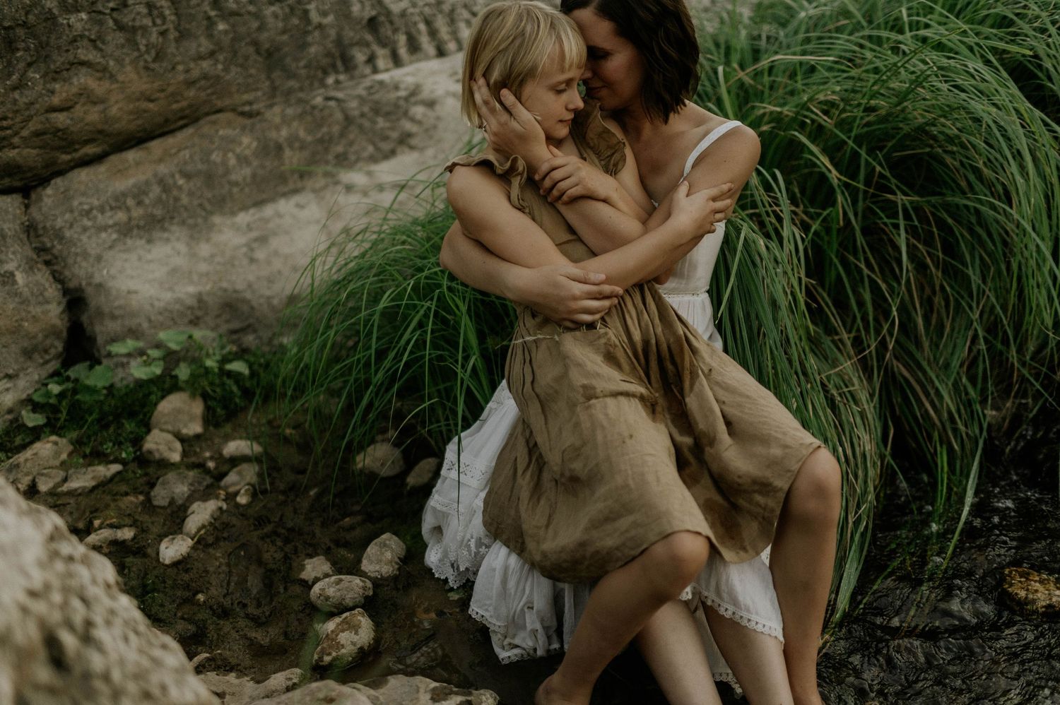 Mother lovingly holding her young blonde daughter among rocky terrain with lush green grass.