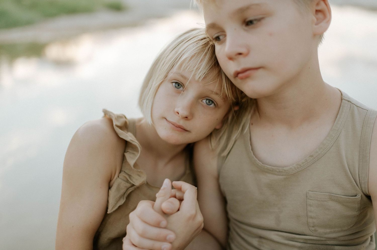 Two blonde siblings holding hands and leaning together near a reflective lake.