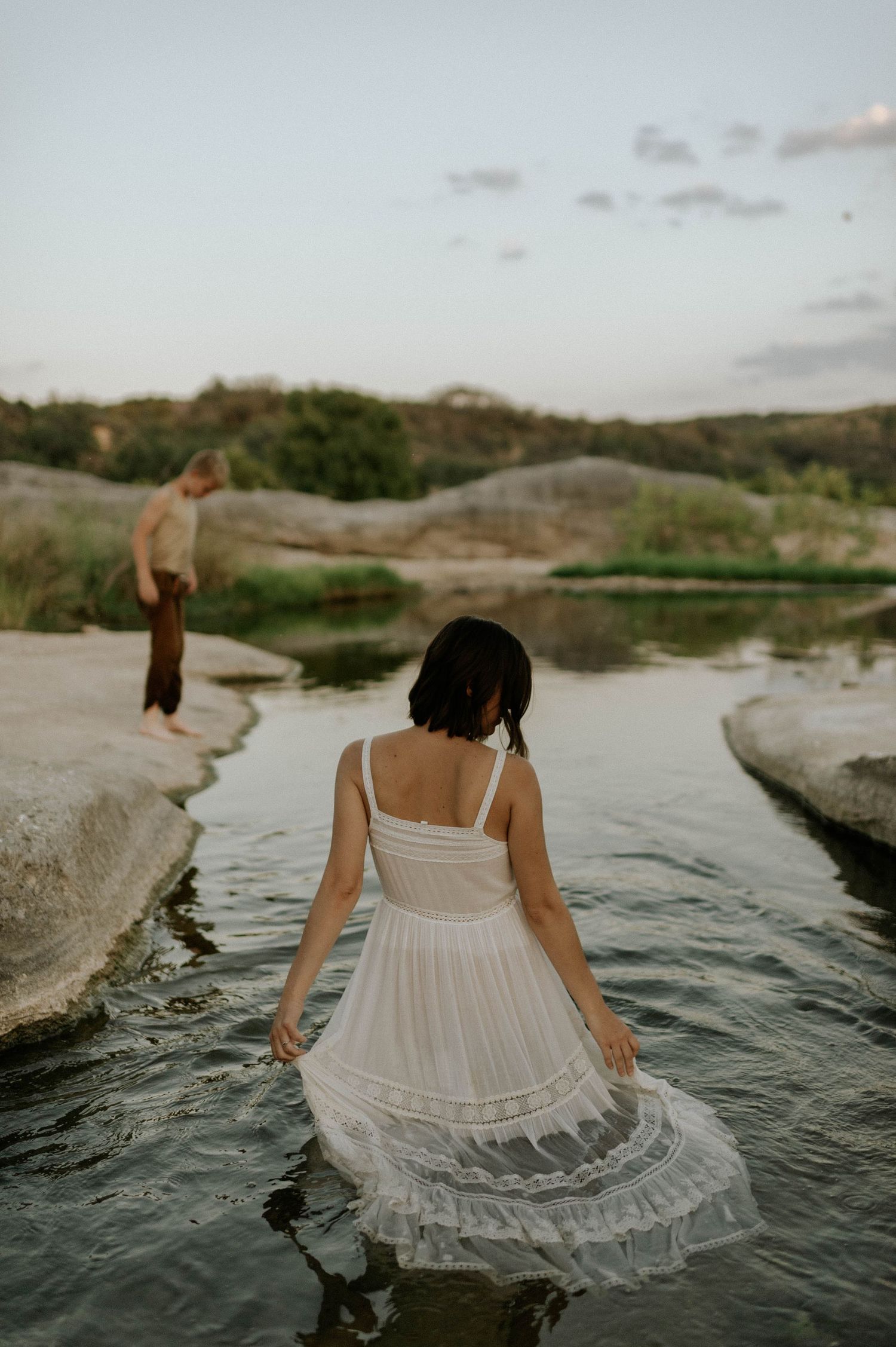 Woman in white dress wading in shallow river with man standing on rocks in background at dusk.