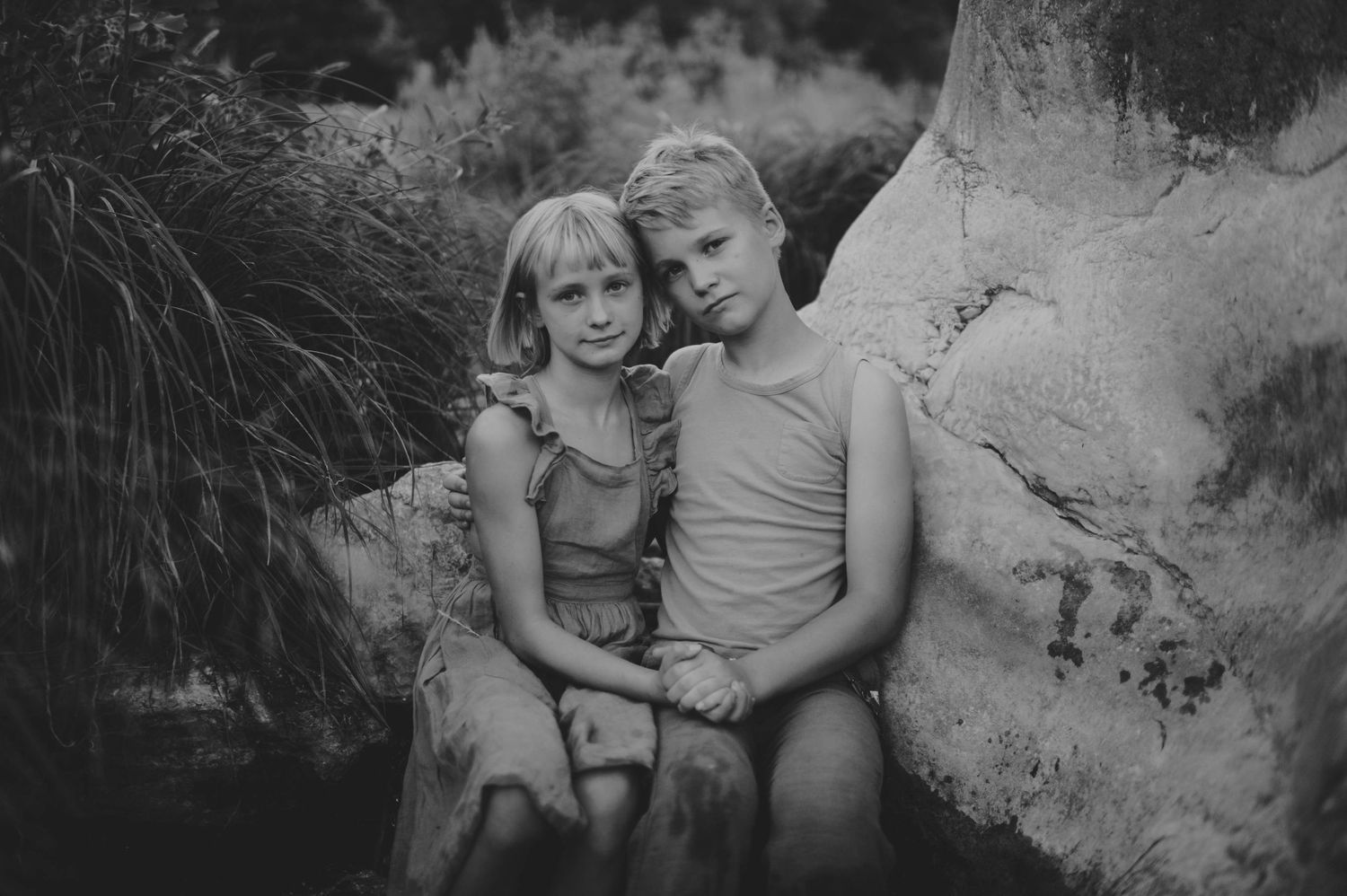 A black and white portrait of two young blonde siblings sitting close together outdoors.