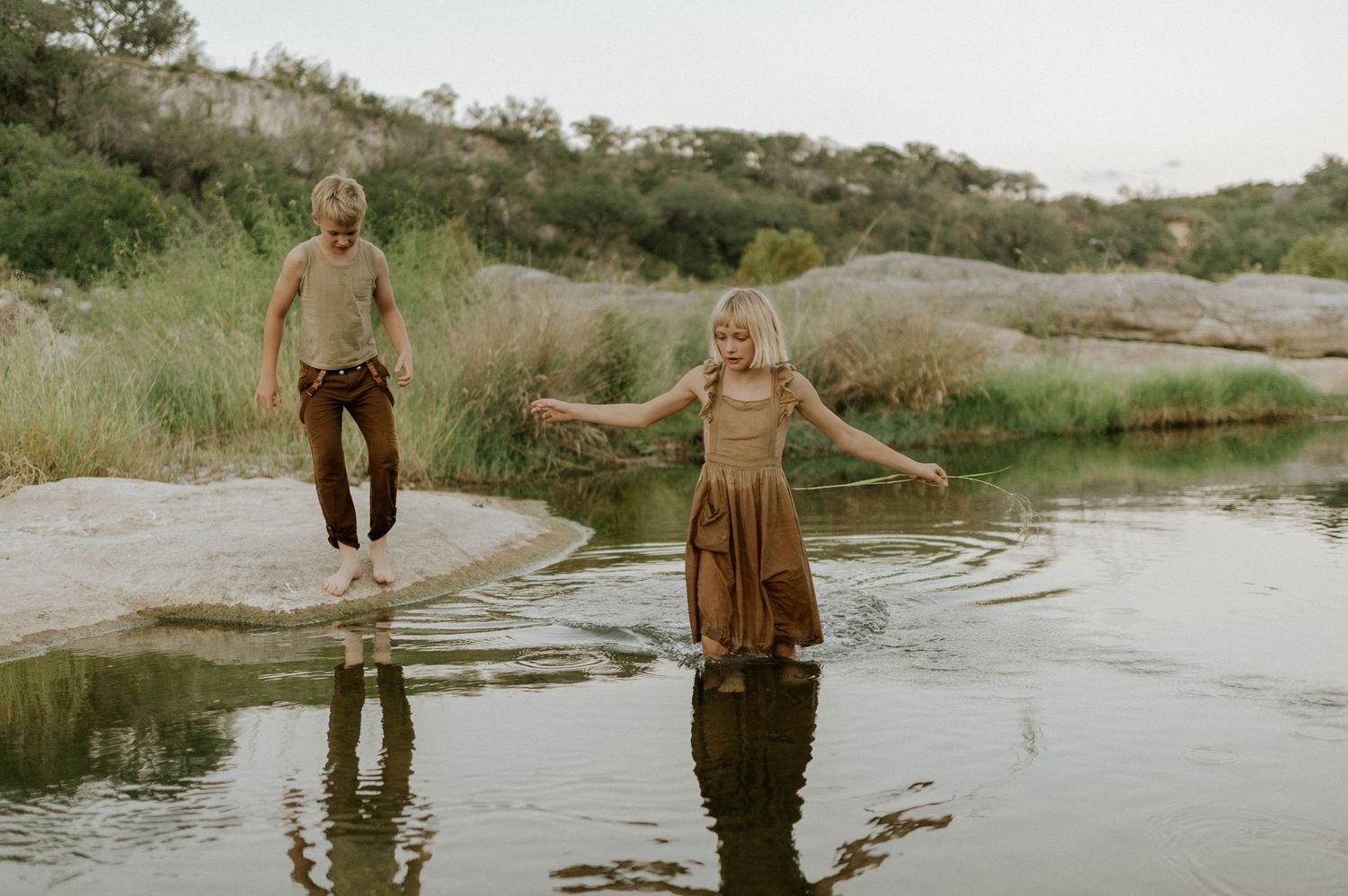 Two children holding hands while wading through shallow river water in a scenic natural setting.