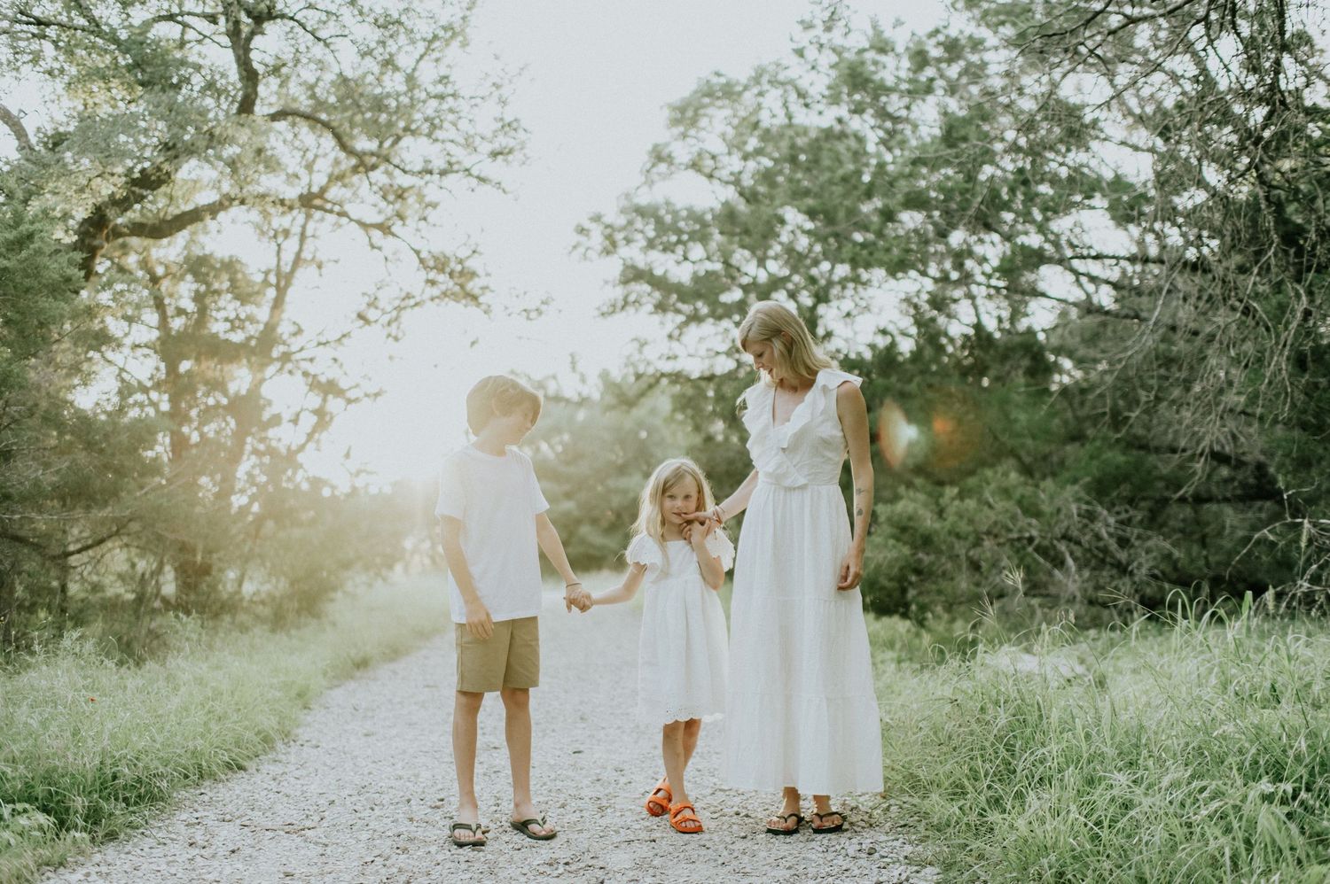 A mother in a white dress holds hands with her two young children on a sunlit gravel path surrounded by lush green trees.